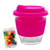 A reusable glass coffee cup with a bright pink silicone lid and sleeve, alongside a packet of multi-coloured jelly beans.
