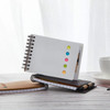A white spiral-bound note pad with colourful sticky dots, placed on a wooden table alongside two other notebooks.