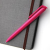 A pink plastic pen rests on a grey textured notebook, featuring a clip and smooth barrel design.