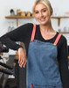 A denim bib apron with red and white striped straps, worn by a model in a café setting.