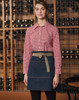 A model wears a denim half apron with a beige waistband, paired with a red checkered shirt against a wine rack backdrop.