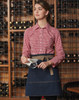 A denim half apron with a light brown belt, worn by a woman in a checked shirt, set against a wine rack background.
