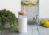 A drink bottle with a white body and bamboo lid, positioned next to a glass dispenser with citrus fruits and greenery.