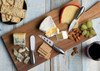 A wooden cheese board featuring various cheeses, crackers, grapes, almonds, and a set of six silver cheese knives.