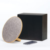 A round, wooden Bluetooth speaker with a grey fabric cover, next to a black box and a smartphone.