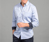 A light blue button-up shirt with long sleeves and a single chest pocket, featuring dark blue cuffs and a logo.