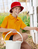A red bucket hat sits on a boy wearing an orange polo shirt, holding a white bucket in a garden setting.