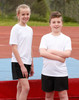 Two children wearing white short sleeve t-shirts and black shorts stand on a sports field. One child has their arms crossed.