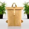 A tan kraft paper backpack with a roll-top closure and adjustable straps, surrounded by potted plants.