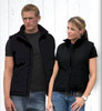 A black men's vest and a black women's vest displayed on models against a white wooden background. Both vests feature zip closures and pockets.