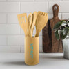 A bamboo cutlery set consisting of various utensils, displayed in a cylindrical holder, on a countertop with a plant and cutting board.