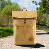 A yellow kraft paper laptop backpack with a fold-over top and front zip pocket, set against a scenic outdoor path.