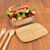 A metal lunch box filled with a sandwich, lettuce, strawberries, and a cutting board made of wood beside it.