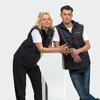 A black unisex puffer vest is worn by two models, one leaning against a clear display stand, showcasing its design.