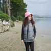 A young woman stands by a lakeside wearing a grey puffer vest and a pink bucket hat among trees.