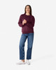 A woman stands wearing a burgundy Gildan Heavy Blend adult crewneck sweatshirt and blue jeans. The sweatshirt features a logo.