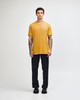 A unisex short sleeve t-shirt in mustard yellow, worn with black cargo pants, featuring a logo.