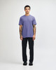 A unisex short sleeve t-shirt in purple, worn by a model with black pants and sneakers, featuring a logo.