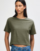 A woman in an olive green boxy T-shirt, styled with high-waisted jeans and curly hair. The shirt has a logo.
