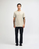 A beige unisex Henley T-shirt featured on a model paired with light blue jeans and black sneakers. The shirt has a logo.