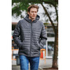 A man wearing a dark grey quilted hoody, walking outdoors surrounded by greenery. The hoody features a logo.