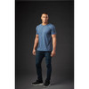 A man models a blue crew neck T-shirt and dark jeans, standing against a dark background. The shirt features a logo.