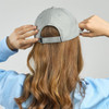 A cap in light grey, featuring an adjustable strap, being worn by a person with long, wavy hair from the back.