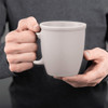 A ceramic mug in a light grey colour, held by a person with two hands. The mug has a smooth finish and a handle.