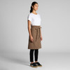 A model wearing a brown canvas half apron with a waist tie, paired with a white t-shirt and black pants.