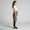 A brown canvas half apron is worn by a person in a white shirt and black pants, featuring a tied waist.
