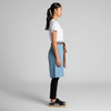 A denim half apron in light blue, featuring a brown tie at the waist, displayed on a model in profile.