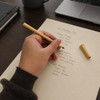 A hand holding a tan-coloured inkless pen, writing on a cream coloured notepad, with a coffee mug and laptop in the background.