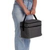 A grey cooler bag with a handle is being held by a person wearing light blue jeans. The bag features a front pocket and branding.