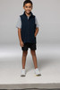 A child models a navy blue vest with a zip, paired with a grey shirt and black shorts. The vest features a logo.