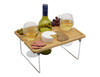 A wooden picnic tray with a bottle of white wine, two glasses, cheese, crackers, olives, and grapes, featuring metallic legs.
