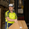 A man wearing a bright yellow and navy polo shirt carries a cardboard box in a warehouse setting.