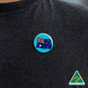 A 25mm button badge featuring the Australian flag, displayed on a dark clothing item, with a logo in the corner.