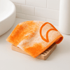 An orange face washer sits on a wooden soap dish, placed next to a white basin.