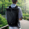 A black roll-up computer backpack with a logo, worn by a person in a checked shirt, against a green forest backdrop.