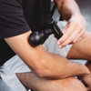 A black mini massage gun being held against a person's arm, showcasing its ergonomic design.