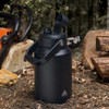 A drink bottle in matte black with a handle and a flip-top spout, set on a forest floor. It features a logo.