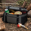 A grey and black cooler bag with drinks, fruits, and food items visible inside, placed among natural foliage.
