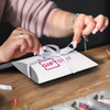 A medium white pillow box with a logo, being tied with a ribbon by a person at a wooden table.