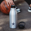 A drink bottle in silver with a black lid and a star logo, set on a textured surface beside a basketball.
