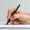 A black metal pen held in a hand, poised above a blank sheet of paper. It features a sleek design and a logo.