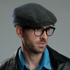 A flat cap in a herringbone pattern, predominantly grey, worn by a model with glasses. Features a logo.