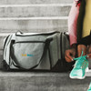 A grey and black duffle bag sits on a concrete surface, with a logo, beside a person tying their shoe.