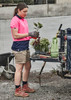A woman in a pink and navy polo shirt wears beige cargo shorts, holding plants beside gardening tools and a wheelbarrow.