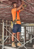 A worker in an orange shirt and navy cargo shorts uses a power tool on a construction site.