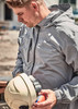 A man in a light grey rain jacket holds a safety helmet and looks down. The jacket features a logo.